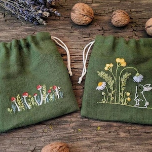 以下が含まれることがあります： 刺繍デザインの緑色の巾着袋が2つ。1つはキノコ、もう1つは花とウサギが特徴です。バッグには白い紐が付いており、クルミとラベンダーのある素朴な木製の背景に置かれています。