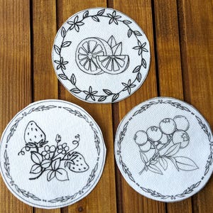 May include: Three white, circular fabric patches with black line drawings of fruit. One features a lemon, another strawberries, and the third blueberries. Each design is framed by a decorative border.