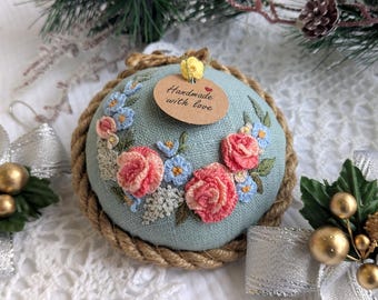 May include: A round, embroidered ornament with a light blue background, featuring pink roses and blue flowers. The ornament is framed with a rope-like border and includes a tag that reads "Handmade with love."