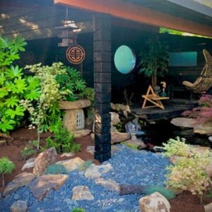 May include: A small, stone Japanese garden lantern with a wooden roof sits in a zen garden with blue gravel, green plants, and a small waterfall.