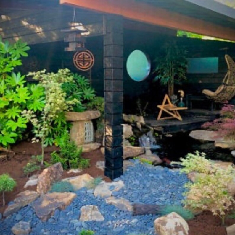 May include: A small, stone Japanese garden lantern with a wooden roof sits in a zen garden with blue gravel, green plants, and a small waterfall.