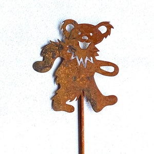 May include: A rusty brown metal bear cutout on a wooden stick. The bear has a smiling face and is in a walking pose. The bear's design is reminiscent of a dancing bear.