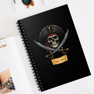 May include: A black spiral notebook featuring a pirate skull and crossbones design. The skull is wearing a black and red pirate hat and the text "Capt. Shells" is written on a scroll.