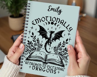 May include: A light blue spiral notebook with the text "Emotionally Attached to Fictional Dragons" and a black silhouette of a dragon with wings spread. The notebook has the name "Emily" written at the top.