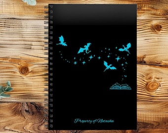 May include: Black spiral-bound notebook with a teal dragon and star design. The design includes flying dragons, stars, and an open book. The text "Property of Natasha" is at the bottom of the notebook.