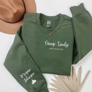 Puede incluir: Una sudadera verde oscuro con el texto "Camp Emily Lake Tahoe 2025" en escritura blanca. La manga presenta el nombre "Megan Johnson" y un gráfico de árbol. También se ven un collar dorado y un sombrero marrón.