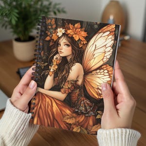 Puede incluir: Una libreta de espiral con una ilustración colorida de un hada con cabello castaño y alas. El hada lleva un vestido adornado con hojas de otoño y flores. El fondo es un bosque oscuro.