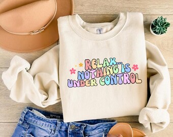 Op de afbeelding: Een crèmekleurig sweatshirt met de tekst "Relax... Nothing is Under Control" in regenboogkleuren met kleine bloem accenten.