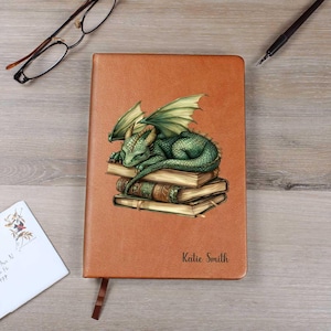 May include: A brown leather journal with a green dragon illustration resting on a stack of books. The dragon has green wings and scales. The journal has a brown ribbon bookmark and the name "Katie Smith" printed on the cover.