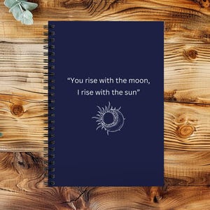May include: A dark blue notebook with a spiral binding. The cover features a white sun and moon design with the quote "You rise with the moon, I rise with the sun."