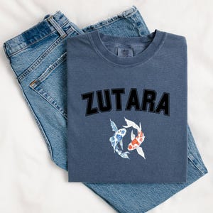 May include: A navy blue t-shirt with the word "ZUTARA" printed in black block letters. Two koi fish, one blue and white and one orange and white, are swimming below the text.