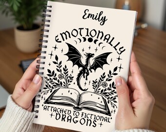 May include: A cream-colored spiral notebook with the text "Emotionally Attached to Fictional Dragons" and a black illustration of a dragon flying above an open book. The name "Emily" is written at the top of the notebook.