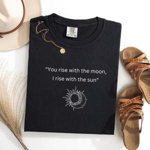 May include: Black t-shirt with a white graphic of a sun and moon with the text "You rise with the moon, I rise with the sun".