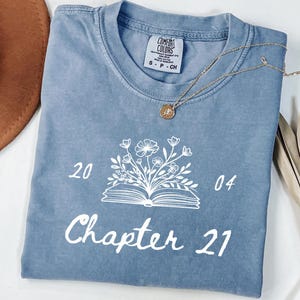 May include: A light blue t-shirt with a white graphic of a book with flowers growing out of it. The text "Chapter 21" is printed below the book. The numbers "20" and "04" are printed on either side of the book.