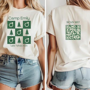 May include: A white t-shirt with a green design featuring a ring, pine trees, and the text "Camp Emily, Lake Tahoe 2025". The back of the shirt has the text "SCAN ME!" and a black and white QR code.