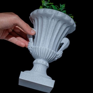 May include: A white ceramic urn with a square base and ornate details. The urn is holding a small green plant.