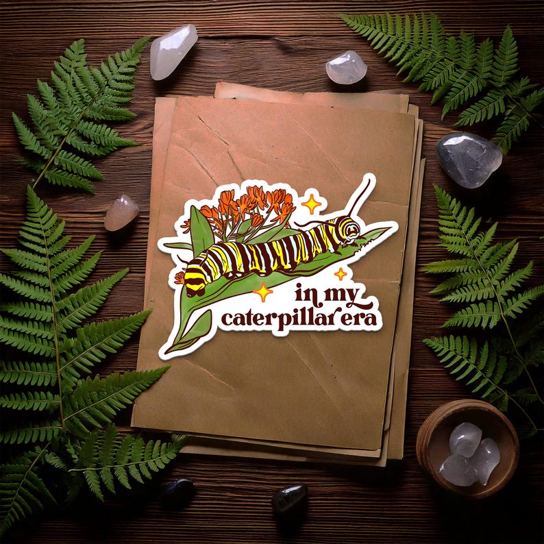 May include: A sticker featuring a caterpillar with yellow and black stripes on a green leaf, with orange flowers and the text "in my caterpillar era". The sticker is on a stack of brown paper, surrounded by ferns and crystals.
