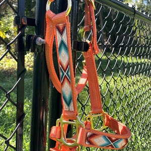 May include: An orange nylon horse halter featuring a decorative woven overlay with a geometric diamond pattern in shades of teal, white, and brown. The halter includes gold-toned metal hardware, including rings and buckles for adjustment.