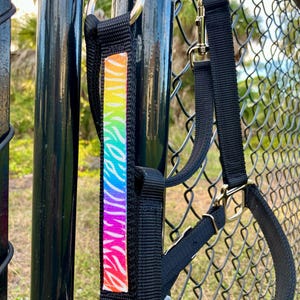 Op de afbeelding: Zwart hondentuig met een regenboog zebra streep accent. Het tuig heeft metalen hardware en een stevig ontwerp. De kleurrijke streep voegt een levendige touch toe aan het functionele huisdieraccessoire.