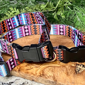 May include: Two dog collars with a colorful geometric pattern in orange, purple, pink, and blue. The collars have black plastic buckles and metal D-rings. The collars are displayed on a wooden surface.