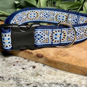 May include: A navy blue dog collar with a decorative pattern in light blue, gold, and white. The collar has a black plastic buckle and a silver metal D-ring for leash attachment. The design is geometric and symmetrical.