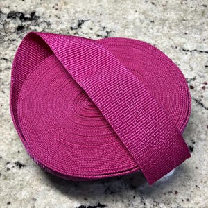 May include: A roll of vibrant fuchsia webbing, with a textured, woven appearance. The webbing is neatly coiled, with a length extending from the roll. This durable material is suitable for various crafting and DIY projects.