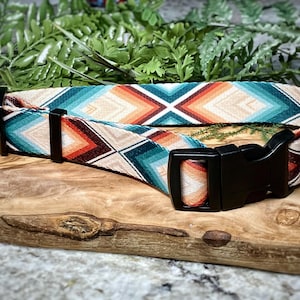 May include: A patterned dog collar with a black plastic buckle and metal hardware. The collar features a geometric design in shades of teal, orange, and cream. The collar is displayed on a wooden surface with greenery in the background.