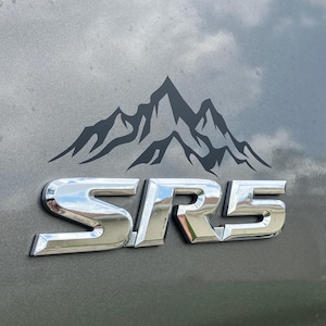 Op de afbeelding: Een zwarte bergketen grafiek boven de chromen letters "SR5" op een grijze ondergrond. De afbeelding is een close-up, die de details van het embleem toont.