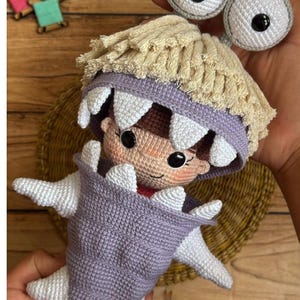 Puede incluir: Muñeca de ganchillo hecha a mano con un personaje con ojos grandes, redondos y grises y una cara marrón claro. La muñeca tiene un cuerpo morado con detalles blancos y una franja beige alrededor de la cabeza. La muñeca se sostiene en una mano.