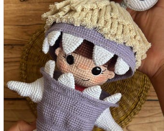Könnte beinhalten: Handgemachte Häkelpuppe mit einer Figur mit großen, grauen, runden Augen und einem hellbraunen Gesicht. Die Puppe hat einen lila Körper mit weißen Akzenten und einen beigen Fransen am Kopf. Die Puppe wird in einer Hand gehalten.