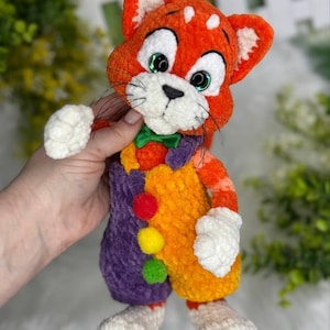 Può includere: Una bambola a forma di gatto all'uncinetto arancione e bianca fatta a mano con occhi verdi, un papillon verde e un abito colorato. La bambola a forma di gatto ha un muso bianco, baffi e zampe bianche. L'abito della bambola è viola, arancione e giallo con pompon colorati.