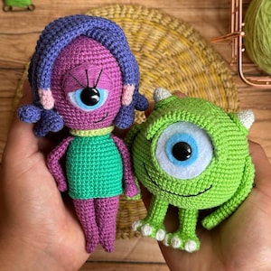 May include: Two handmade crochet dolls, one purple and one green. The purple doll has a single eye, blue hair, and a green top. The green doll is a one-eyed monster with small horns and white feet. Both dolls are held in hands.