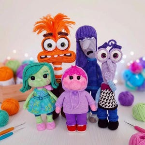Puede incluir: Una colección de cinco muñecos de ganchillo hechos a mano, cada uno representando un personaje de la película animada "Del revés". Los muñecos presentan colores vibrantes, incluyendo verde, naranja, morado y rosa, con expresiones faciales detalladas y atuendos únicos. Los muñecos se exhiben sobre una superficie blanca.