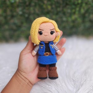 May include: A crochet doll featuring a character with short yellow hair, black eyes, and pink cheeks. The doll wears a blue vest over a black shirt with white and black striped sleeves, a blue skirt, a brown belt with a gold buckle, and brown boots.