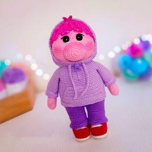 以下が含まれることがあります： ピンクの顔、鼻、髪の毛を持つ手作りの編み人形。紫色のパーカー、ズボン、赤い靴を履いています。人形は黒い目を持ち、柔らかい糸で作られています。ユニークな手作りのアイテムです。