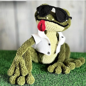 Pode incluir: Um sapo verde de crochê feito à mão usando óculos de sol pretos e um colete branco. O sapo tem uma língua vermelha saindo e está sentado na grama verde. Os braços e pernas do sapo estão estendidos.