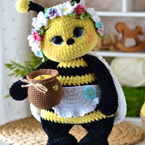 May include: A crocheted bee doll with yellow and black stripes, wearing a white apron with a blue flower and a crown of colorful flowers. The doll has large blue eyes with long lashes and holds a small brown crocheted honey pot with a twine tie.