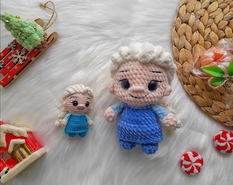 May include: Two crocheted dolls resembling a character with blonde hair and a blue dress. One doll is larger than the other. The dolls are on a white, fluffy surface, with a red and green Christmas ornament and two red and white candies.