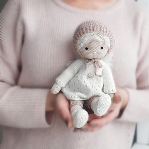 May include: A handmade crochet doll with a white dress, matching socks, and a light brown hat. The doll has white hair and a light brown bow around its neck. The doll is being held in someone's hands.