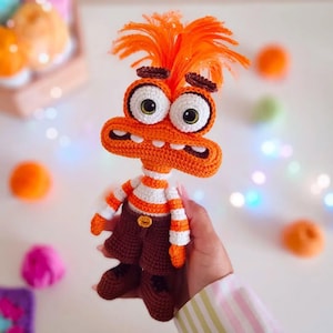 Puede incluir: Muñeco monstruo naranja tejido a crochet, con grandes ojos con borde blanco y boca ancha. El muñeco lleva una camisa a rayas, pantalones cortos marrones y zapatos. Un juguete hecho a mano único.