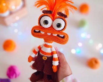 Könnte beinhalten: Gehäkelte orangefarbene Monsterpuppe mit großen, weiß umrandeten Augen und einem breiten Mund. Die Puppe trägt ein gestreiftes Hemd, braune Shorts und Schuhe. Ein einzigartiges handgemachtes Spielzeug.