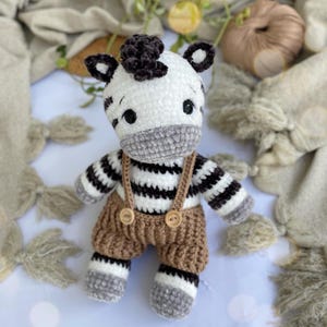 Op de afbeelding: Handgemaakte gehaakte zebra pop met een witte body, zwarte strepen en een grijze snuit. De zebra draagt een bruine tuinbroek met houten knoopdetails. De pop heeft zwarte ogen en een donkerbruine manen.