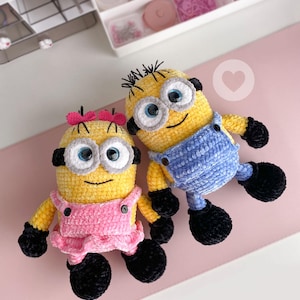Puede incluir: Dos peluches de ganchillo con forma de personajes amarillos, ojos grandes y redondos, guantes negros y zapatos negros. Uno lleva un peto rosa con falda y lazo, el otro un peto azul. Ambos tienen cuerpos hechos de hilo con textura.