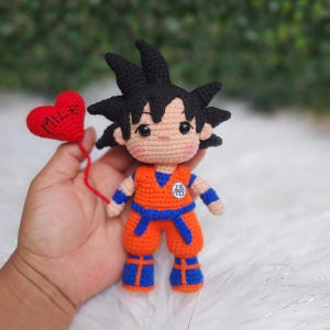 May include: A crocheted doll of a character with spiky black hair, wearing an orange gi with a blue belt, blue wristbands, and blue boots. The doll holds a red heart-shaped balloon with the word MILK embroidered in black thread on the front.