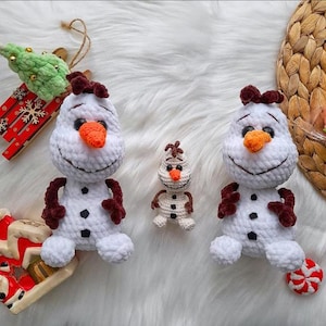 May include: Crocheted snowman toys with orange carrot noses, black button eyes, and brown twig arms. The snowmen have a smiling face and are surrounded by Christmas decorations, including a red and white sled and a green Christmas tree.