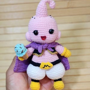 Könnte beinhalten: Eine gehäkelte rosa Puppe mit einer gekringelten Antenne, schwarzen Augen und einem kleinen roten Mund. Die Puppe trägt einen violetten Umhang, eine schwarze Weste mit Goldrand, eine weiße Hose und gelbe Stiefel. Sie hält eine kleine blaue Eistüte.
