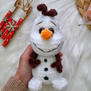 K&ouml;nnte beinhalten: Ein handgefertigtes wei&szlig;es Schneemann-Pl&uuml;schtier mit braunen Haaren, Armen und schwarzen Kn&ouml;pfen. Der Schneemann hat eine orangefarbene Karottennase und ein l&auml;chelndes Gesicht. Ein winterliches Thema.