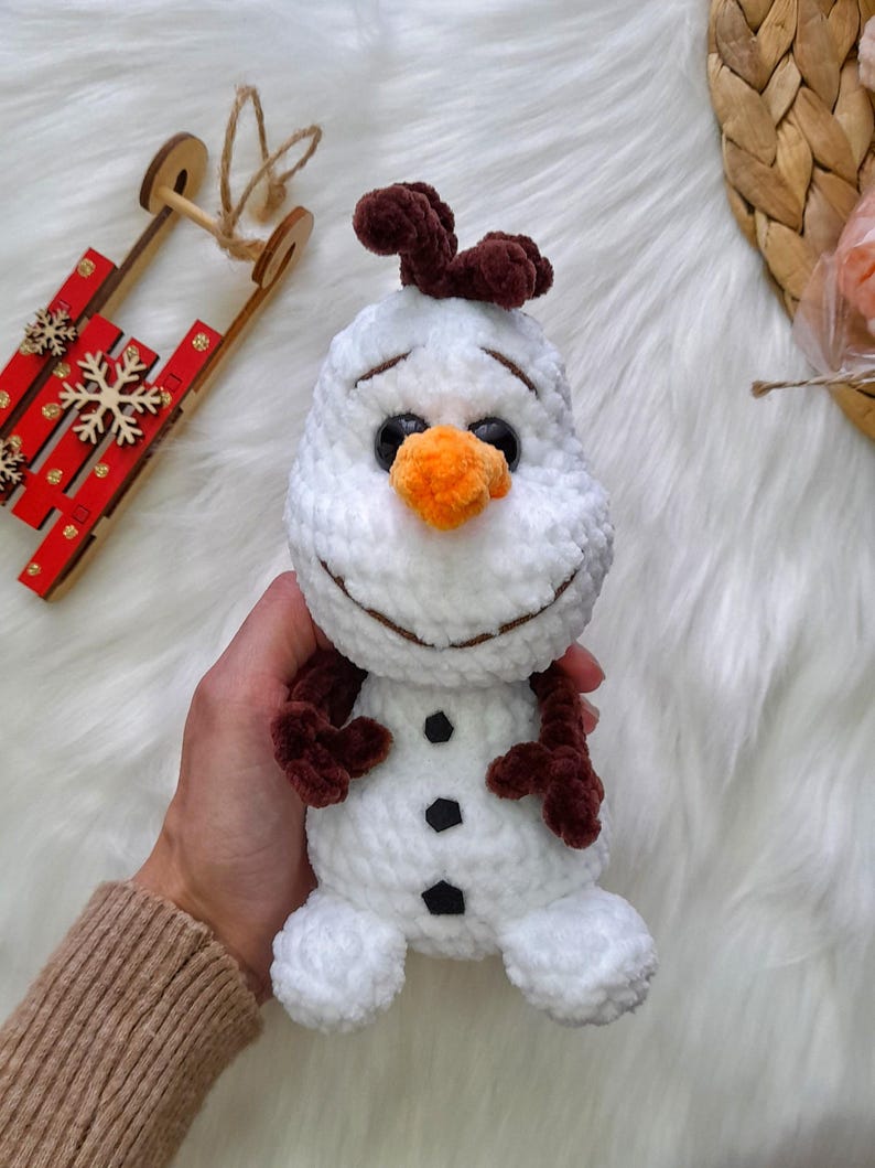 K&ouml;nnte beinhalten: Ein handgefertigtes wei&szlig;es Schneemann-Pl&uuml;schtier mit braunen Haaren, Armen und schwarzen Kn&ouml;pfen. Der Schneemann hat eine orangefarbene Karottennase und ein l&auml;chelndes Gesicht. Ein winterliches Thema.