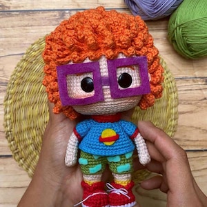Könnte beinhalten: Handgemachte Häkelpuppe mit orangefarbenem Haar, lila Brille und einem blauen Hemd mit gelbem Planetenmuster. Die Puppe trägt grüne Shorts, rote Schuhe und wird in den Händen einer Person gehalten.