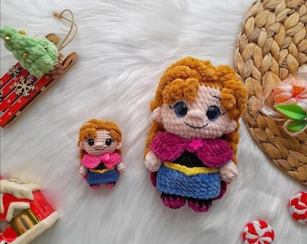 Könnte beinhalten: Zwei Häkelpuppen, eine große und eine kleine, die einer Figur mit orangefarbenem Haar, einem rosa Umhang und einem blauen Kleid ähneln. Die Puppen befinden sich auf einer weißen, flauschigen Oberfläche, mit einem roten und grünen Weihnachtsschlitten und einem geflochtenen Korb im Hintergrund.